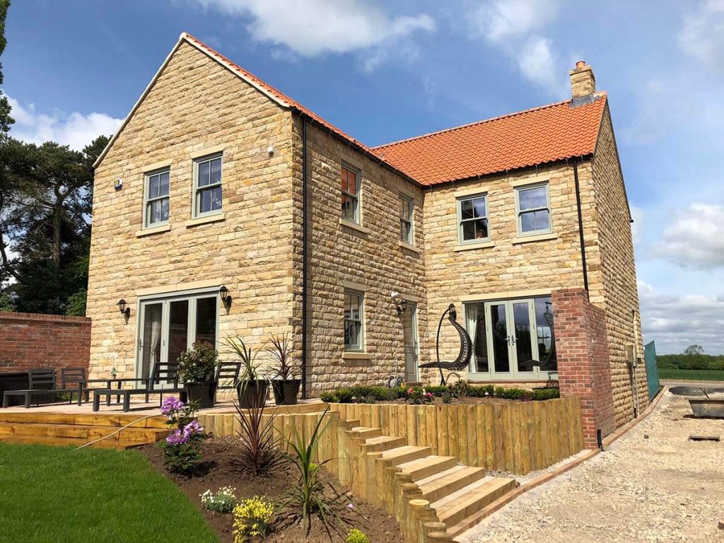 An elegant sandstone brick house with a striking red roof, featuring beautifully crafted Chartwell green windows and patio doors. The residence exudes a charming and inviting aura, showcasing a perfect blend of natural and modern elements.